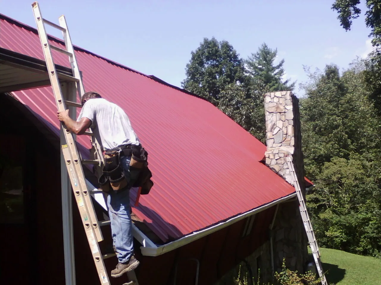 Roofer on a ladder beside a red metal roof and stone chimney — Soffit & Fascia Repair by Roofing Brothers in Sutherlin